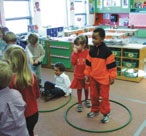 Situation de salle de classe. Il y a deux cerceaux, parterre, dans le premier cerceau, il y a un enfant debout et l’autre est assis. Dans le deuxième cerceau, les deux enfants sont debout.  Il y a d’autres enfants dans la classe.