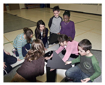 Students are sitting on the floor, manipulating large cardboard shapes.