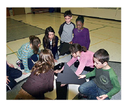 Students are sitting on the floor, manipulating large cardboard shapes.